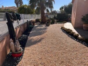Allée de gravier pêche et jardin sec méditerranéen avec grand palmier, cactus en pot rouge et bordures de roches noires. Manteaux sur la clôture.