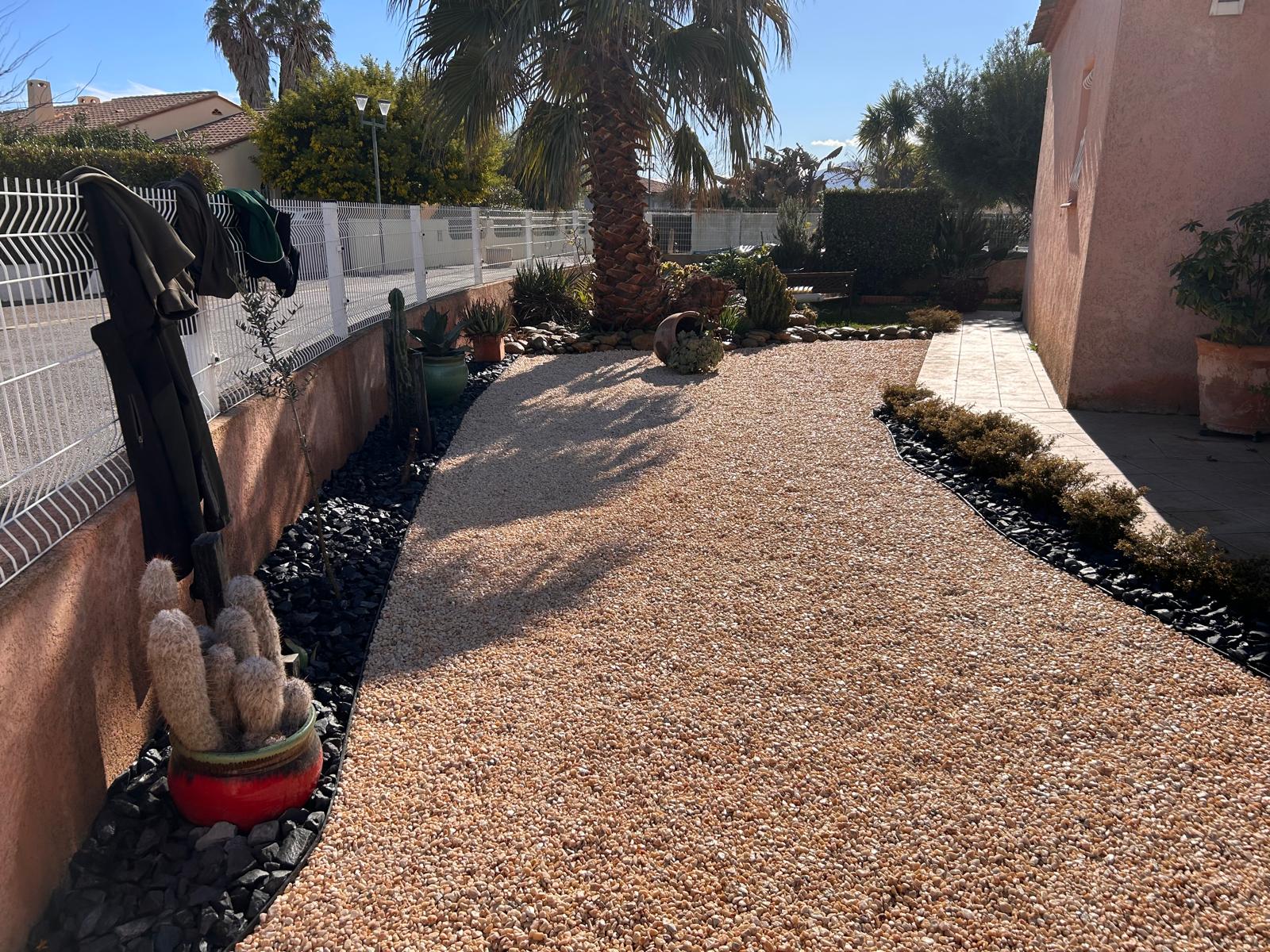 Allée de gravier pêche et jardin sec méditerranéen avec grand palmier, cactus en pot rouge et bordures de roches noires. Manteaux sur la clôture.