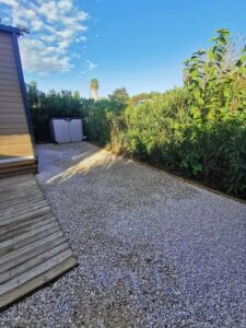 Allée de gravier devant un mobil-home. Terrasse bois usée, abri de jardin gris et haie dense en pleine lumière sous un ciel bleu dégagé.