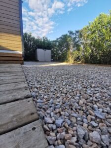Allée de gravier coloré en gros plan, bordée par une terrasse en bois patiné, avec un cabanon gris et une haute haie verte sous un ciel bleu.