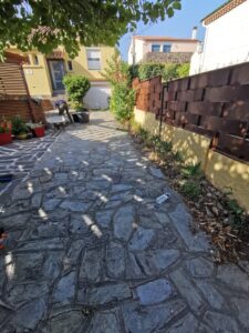 Allée pavée en pierre naturelle sous les arbres, menant à une maison jaune. Un ouvrier vide une brouette pour des travaux de jardinage.