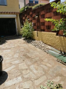 Patio en pierres irrégulières menant à un garage. Bordure de galets et arbuste fleuri devant un mur en métal rouillé.