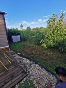 Aménagement de jardin près d'une terrasse en bois. Un homme pose du gravier blanc le long d'un cabanon, entouré d'une végétation dense sous ciel bleu.