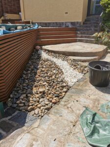 Aménagement paysager d'un ruisseau sec partant d'une jarre, longeant une structure en bois. Galets bruns et bande de galets blancs sur dalles de schiste.