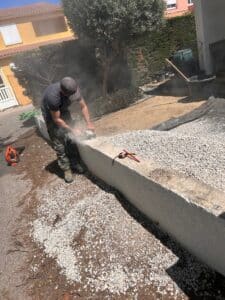 Ouvrier coupant un muret de béton avec une meuleuse, créant un nuage de poussière blanche sur un chantier ensoleillé.