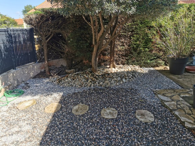 Aménagement d'une allée de jardin moderne avec du gravier noir et des pas japonais, bordée par un massif de galets clairs entourant un arbre.