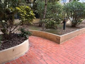 Allée en briques rouges menant à des jardinières surélevées beiges courbées. Végétation dense, paillis d'écorce et lampadaire de jardin.