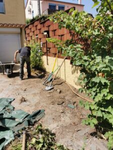 Aménagement extérieur. Un jardinier pelle la terre près d'une clôture en acier Corten texturé et d'un mur jaune, outils au sol.