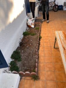 Parterre de jardin en bordure de briques festonnées, sol nu. Patio en carrelage orange et mur blanc crépi. Un sac de terre près des pieds.