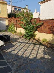 Patio en dalles de schiste sous des ombres. Clôture rouillée Corten, mur jaune et arbustes verts délimitent l'espace.