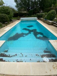 Piscine turquoise extérieure très sale, couverte de feuilles mortes et de débris sur les marches et formant un grand motif au fond. Bâche automatique visible.