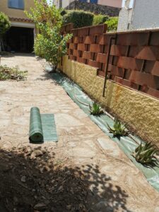 Allée latérale ensoleillée avec pose de toile de paillage verte et plants d'aloès, bordant une clôture métallique rouillée.