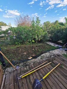 Préparation du jardin. Pelle et râteaux sur une terrasse en bois bordant la terre labourée et les arbustes méditerranéens sous un ciel bleu.