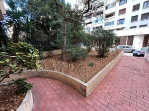 Parterre végétal urbain délimité par une bordure en béton et une clôture de bambou, longeant des pavés rouges menant à une résidence.