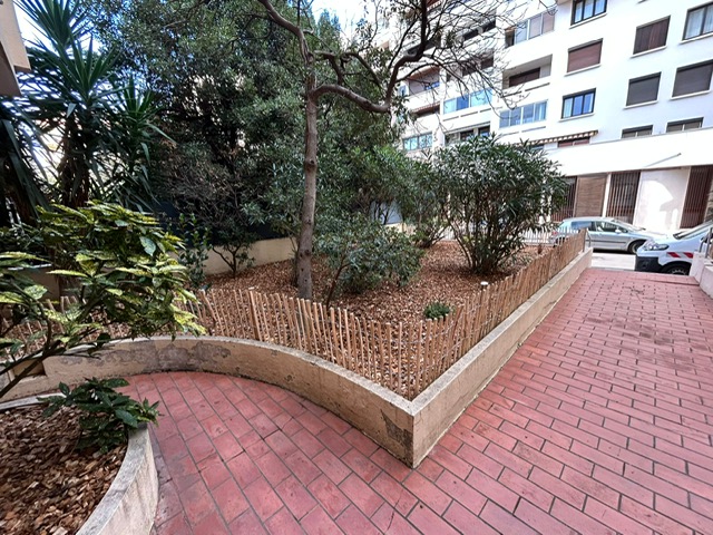 Parterre végétal urbain délimité par une bordure en béton et une clôture de bambou, longeant des pavés rouges menant à une résidence.